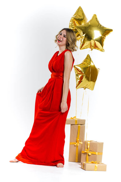 Party photo of elegance sexy lady in red dress with red lips and blond beautiful curly hair, smiling. Background of gold air balloons and gifts. Concept of the sale - Zdjęcie, obraz