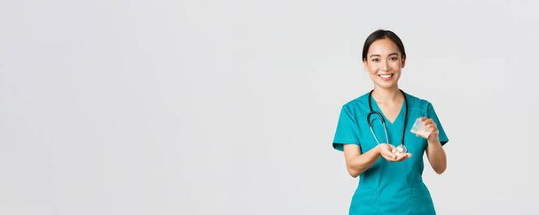 Gezondheidswerkers, het voorkomen van virussen, quarantaine campagne concept. Glimlachende vriendelijke Aziatische vrouwelijke arts, arts in scrubs, geven patiënt medicatie uit container, witte achtergrond - Foto, afbeelding