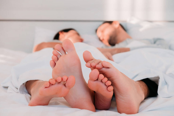 ベッドの下のカバーの下で眠っている男と女の素足のクローズアップ。脂っこい部屋で家族のマシューの朝。人の愛と関係 - 写真・画像