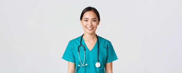 Медицинские работники, профилактика вируса, концепция карантинной кампании. Крупный план улыбающейся приятной азиатской медсестры, врача в форме, выглядящего оптимистично, слушающего пациента, белого фона - Фото, изображение