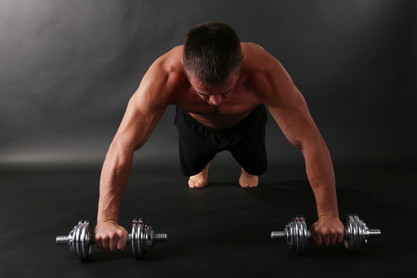 Handsome young muscular sportsman execute exercise with dumbbells on dark background - Photo, image