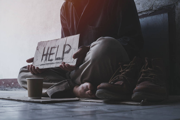 Ein obdachloser erwachsener Mann sitzt auf der Straße im Schatten des Gebäudes und bettelt mit einem Schild um Hilfe und Geld. Obdachlosenkonzept - Foto, Bild