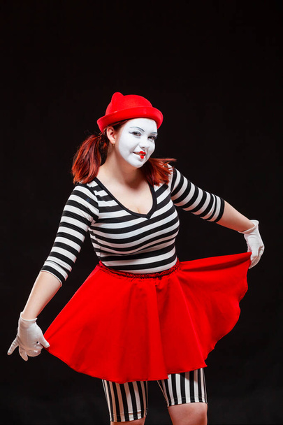 Portrait of female mime artist performing, isolated on black background. Woman in striped clothes holding her red skirt and smiling - Fotó, kép