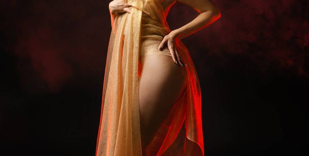 Young nude woman posing in studio with a golden shawl on a dark background - Фото, изображение