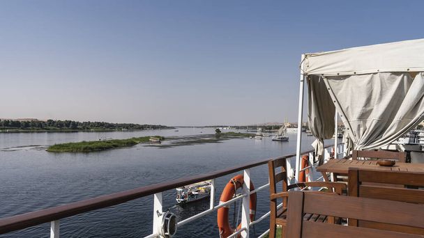 Tavoli e sedie in legno sono installati sul ponte superiore della nave da crociera. Dal ponte si può vedere il fiume Nilo Azzurro, le rive verdi e l'isola.Barche sull'acqua. Cielo azzurro limpido. Egitto - Foto, immagini