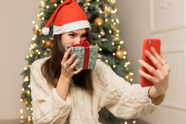 Das junge Mädchen telefoniert mit den Eltern und zeigt ein grün-rotes Weihnachtsgeschenk. Glücklich lächelnde Frau mit rotem Hut auf dem Kopf sitzt neben einem festlichen Baum - Foto, Bild