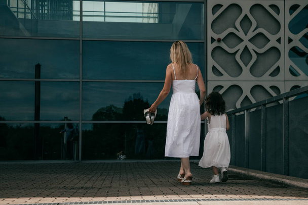 Κοριτσάκι με τη μητέρα της ντυμένο με λευκά βαμβακερά ηλιοβασιλέματα στο φόντο του όμορφου κτιρίου της πόλης την καλοκαιρινή μέρα. Ευτυχισμένη σχέση. Όμορφοι άνθρωποι. Χαρά. Φυσικά υλικά. Πίσω όψη - Φωτογραφία, εικόνα
