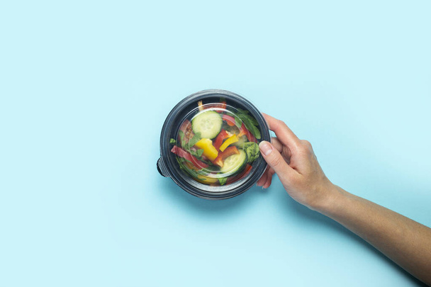 Женская рука держит закрытую пластиковую одноразовую тарелку с овощным салатом на светло-голубом фоне. Вид сверху, плоский. - Фото, изображение