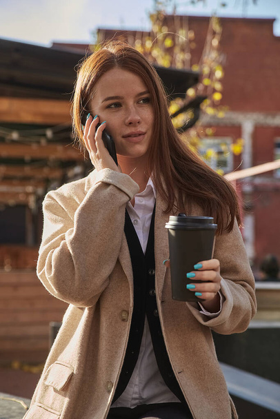 donna caucasica in elegante cappotto beige chiamata da smartphone, bere caffè - Foto, immagini