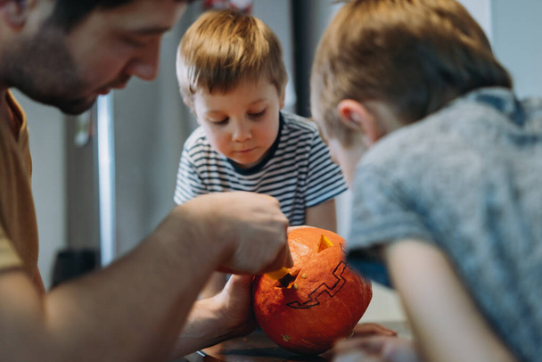 preparing for halloween celebration. Caucasian man withhis cute sons carving out eyes on a pumpkin to make traditional Jack lantern. Image with selective focus. - Foto, Imagem