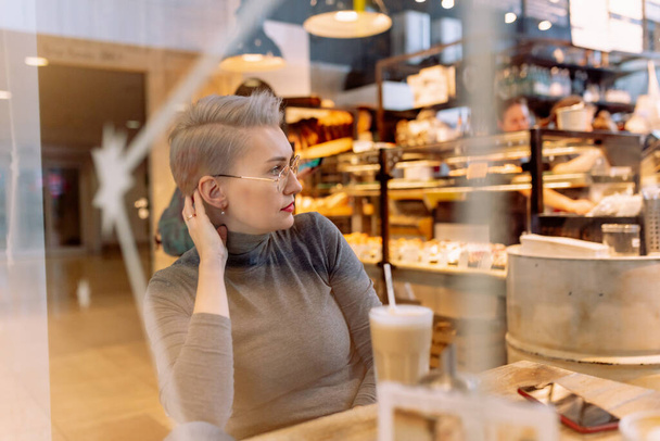 young caucasian blond woman recording audio message with smartphone sitting in cafe. View through glass. Image with selective focus and noise effect - Фото, зображення