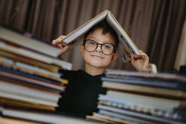 cute caucasian boy wearing glasses covering head with book. Cheerful boy peeking from behind piles of books. Image with selective focus and noise effect - Foto, imagen