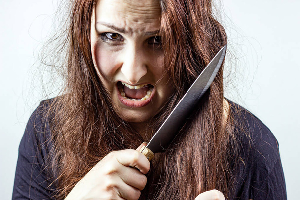Junge kaukasische wahnsinnig gestresste brünette Frau mit Messer in schwarz vor hellem Hintergrund. Halloween-Konzept. - Foto, Bild