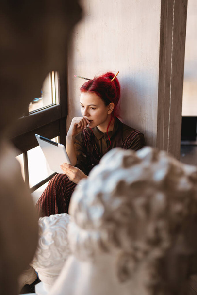 Young woman artist sitting on a windowsill in an art studio holding tablet device - Φωτογραφία, εικόνα