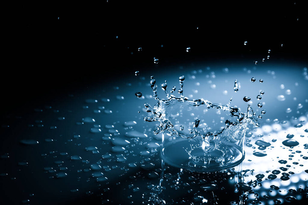 Beautiful splash crown from a drop of water falling on a hard surface. In blue light on a black background - Photo, Image