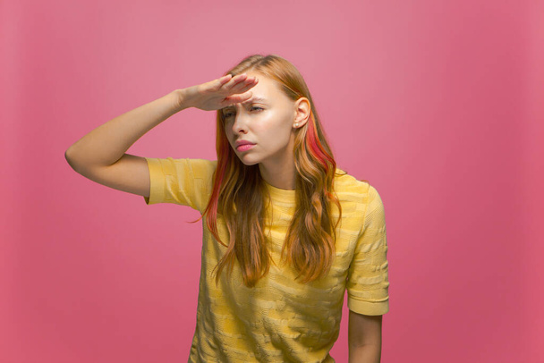 Focused attentive serious young woman looking far away with hand over head, searching on pink studio background - 写真・画像