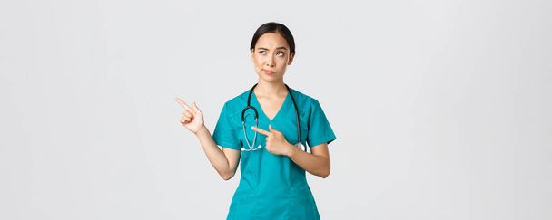 Covid-19, Gesundheitspersonal, Pandemiekonzept. Skeptisch und zweifelhaft hübscher asiatischer Arzt, Krankenschwester im Peeling grinst, zeigt in die linke obere Ecke, blickt mit zögerndem Gesicht, weißer Hintergrund - Foto, Bild