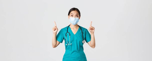 Covid-19, malattia coronavirica, concetto di operatore sanitario. Incuriosito ed eccitato medico donna asiatica, infermiera in maschera medica e guanti, guardando e puntando le dita verso l'alto, sfondo bianco - Foto, immagini