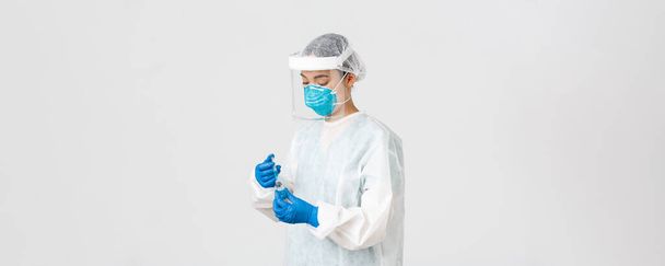 Covid-19, coronavirus disease, healthcare workers concept. Serious-looking female physician, asian doctor in personal protective equipment insert syringe in ampoule with vaccine, white background - Foto, Imagem