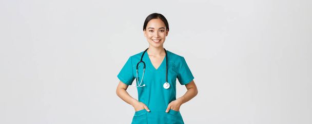 Covid-19, gezondheidswerkers, pandemisch concept. Zelfverzekerde glimlachende mooie Aziatische vrouwelijke arts, arts op zoek naar vastberaden en upbeat, hand in hand in zakken van scrubs, onderzoeken patiënten in de kliniek - Foto, afbeelding