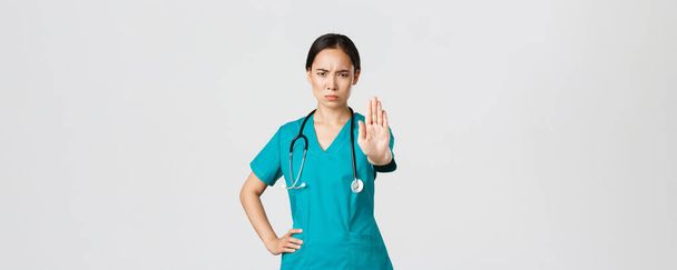 Covid-19, trabajadores de la salud, concepto pandémico. Enojado médico asiático de aspecto serio, médico o enfermera en matorrales frunciendo el ceño disgustado, extender la mano para mostrar parada, en desacuerdo, prohibir o prohibir - Foto, Imagen
