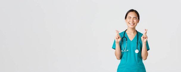 Covid-19 、医療従事者、パンデミックの概念。楽観的な幸せと希望のアジアの女性看護師,昇進を待っているインターン,クロス指幸運と近い目,笑顔興奮 - 写真・画像