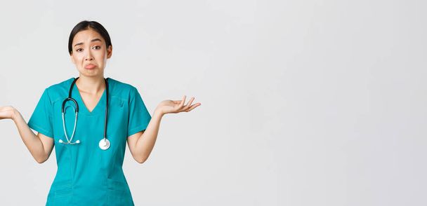 Ковид-19, медицинские работники, пандемия. Портрет невежественной азиатской медсестры, женщина-врач пожимает плечами и раздвигает руки боком, не знаю, не могу помочь, белый фон - Фото, изображение