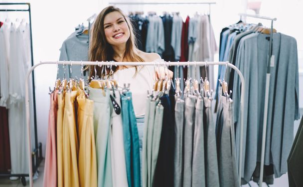 Happy female shopper standing behind clothes rack and smiling while selecting new apparel in modern store and looking at camera - Фото, изображение