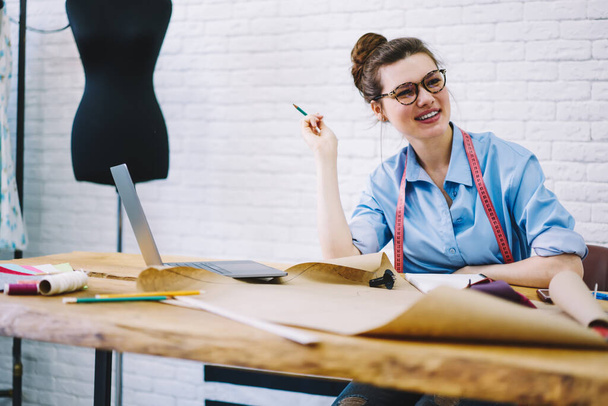 Low angle of cheerful young female tailor in casual wear eyeglasses and measuring tape around neck sitting at wooden table with drawings and laptop in modern workshop - Foto, immagini