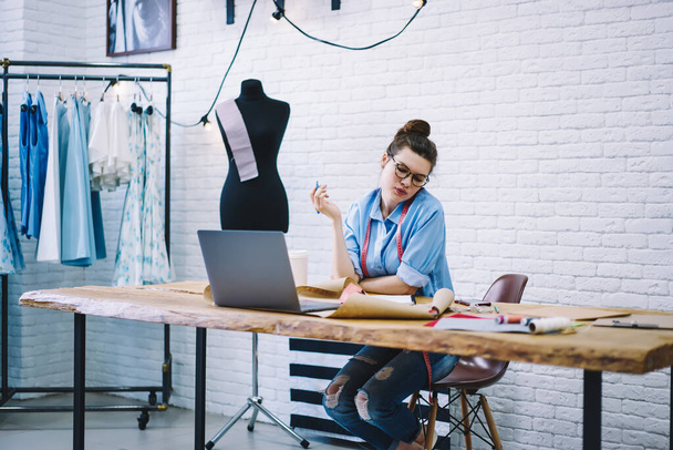 Thoughtful female fashion designer drawing sketches for new collection sitting at table with laptop in studio with mannequin and row of dresses on hangers - Photo, image