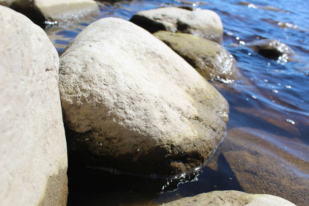 чистая вода течет над большими камнями летом / фото камни и вода. Большие камни лежат в воде. Между камнями чистая вода. отражения солнца отражаются в воде. красивый пейзаж. - Фото, изображение