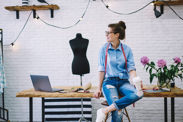 Smiling female dressmaker in glasses with takeaway hot beverage resting after work and thinking about design project in light studio with mannequin - Foto, imagen