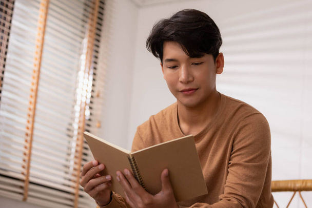 At home, a handsome young Asian businessman in casual clothing reads a book on the couch. - Zdjęcie, obraz