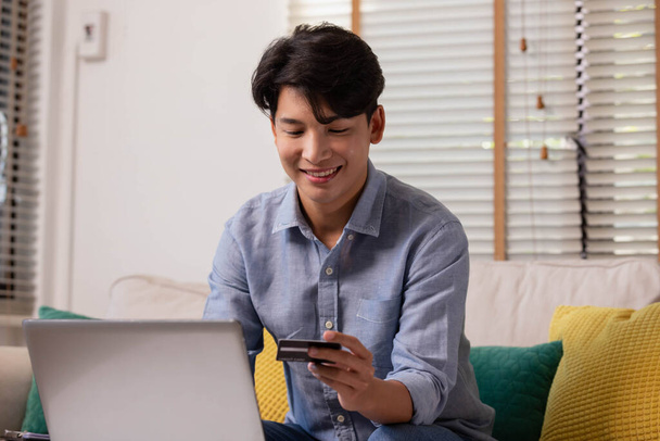 Ázsiai férfi fizet az interneten keresztül. Otthon a kanapén egy vásárló online vásárol hitelkártyával és okostelefonnal. Az internetes vásárlás fogalma. - Fotó, kép