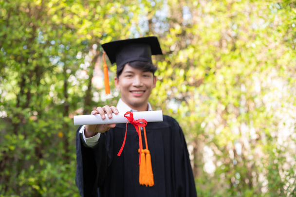 In the graden, a happy young Asian man wears graduation gowns and has a diploma. - Photo, Image