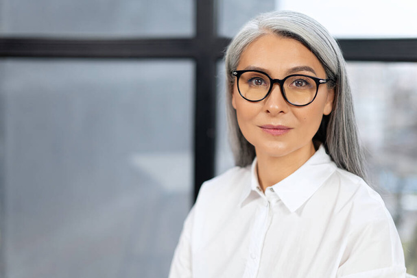 Portrait view of confident and smiling old senior business woman wearing glasses looking at the camera with pleasure smile. Employee worker sitting at the office concept - Фото, зображення