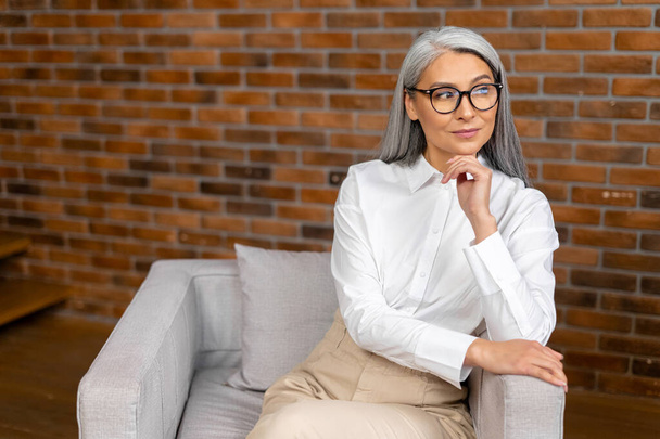 Portrait of an elegant stylish mature middle-aged businesswoman, ceo in glasses sitting in the modern office and posing with the arms folded - Fotó, kép
