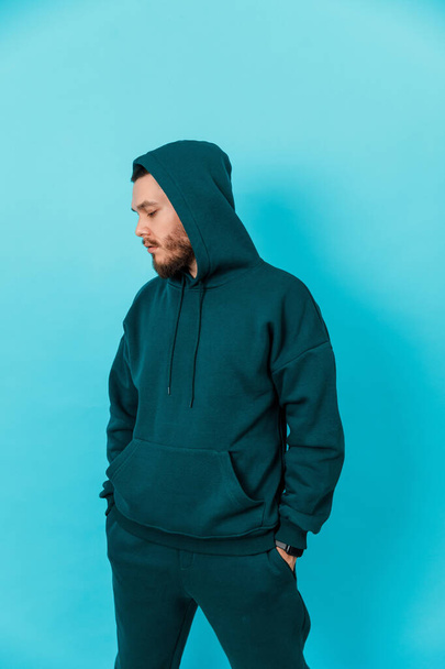 handsome young man with a beard in a green tracksuit looks to the side and holds his hands in his pockets on a blue background. isolated - 写真・画像