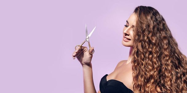 Beautiful woman with long curly hair holds hairdressers scissors, isolated on pink studio background  - Fotó, kép