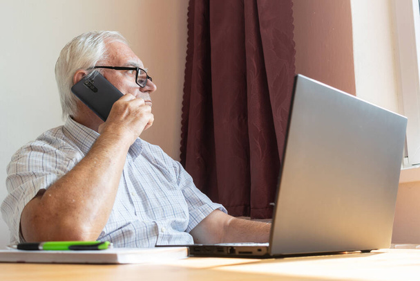 Главная тема не в фокусе, старший мужчина мужчина сидеть рядом за столом, домашний офис онлайн ноутбук ноутбук работы бизнес расстояние фрилансер держать смартфон - Фото, изображение