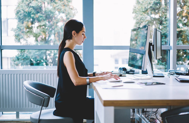 Side view of concentrated woman entrepreneur in black clothes sitting at table and typing on computer while working on project in contemporary office with panoramic windows in daytime - 写真・画像
