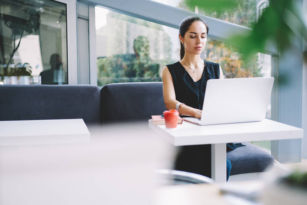 Komoly női szabadúszó alkalmi ruhában nézi a képernyőt az asztalnál ülve elvitelre szánt kávéval, miközben laptopot használ a munkához. - Fotó, kép