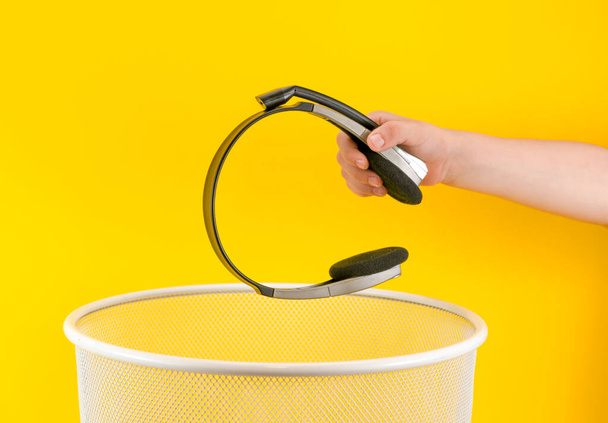 Hand throws stereo headphones into trash bin with package on yellow background - Fotó, kép