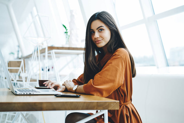 Young positive female freelancer in casual outfit looking at camera sitting at table with smartphone and cup of coffee while working on laptop - Foto, Imagen