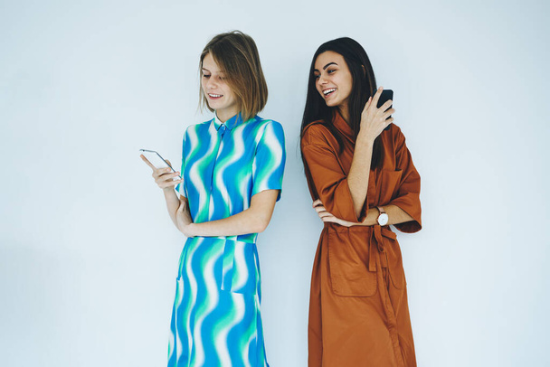 Happy young women wearing trendy dress standing back to back with smartphone in hand and smiling with white wall in background - Фото, изображение