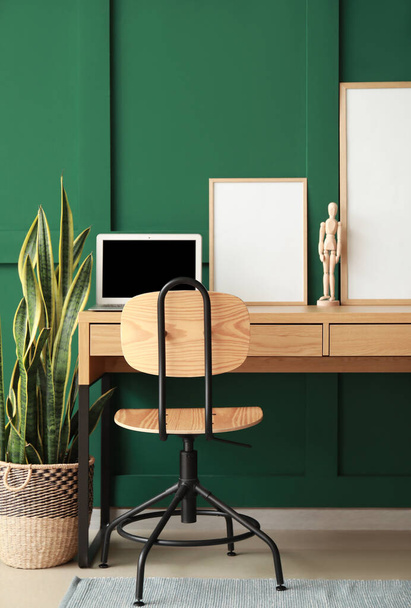 Moderne werkplaats met laptop, etalagepop en lege lijsten in de buurt van groene muur - Foto, afbeelding