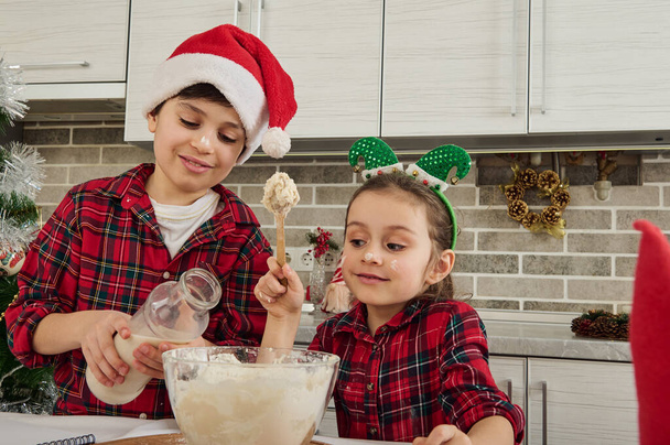 Cheerful brother and sister, cute Caucasian children spend time together at home kitchen. Adorable boy pours milk and baby girl kneads dough for Christmas cookies. Perfect happy family relationships - Photo, Image