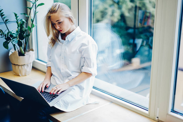 Gericht jonge vrouw in casual witte outfit werken aan nieuw project met behulp van laptop terwijl zitten op vensterbank en kijken naar het scherm - Foto, afbeelding