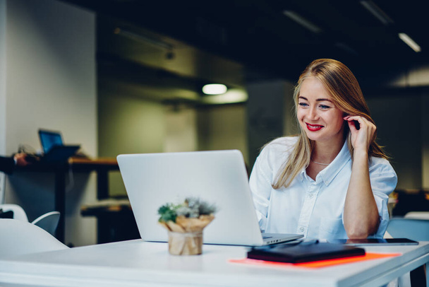 Happy female freelancer dressed in white shirt and with red lipstick using laptop for work while sitting in cafe at table - Фото, изображение