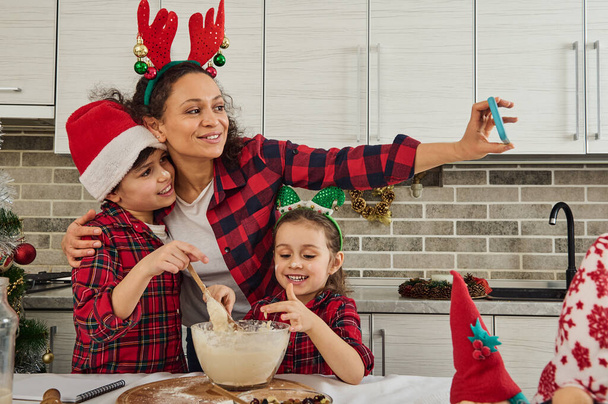 Hermosos niños europeos amasando masa, ayudando a su madre en la cocina de pan de Navidad y galletas. Encantada mamá grabando vídeo y haciendo selfie, abrazando a su hijo y su hija. Familia feliz - Foto, Imagen